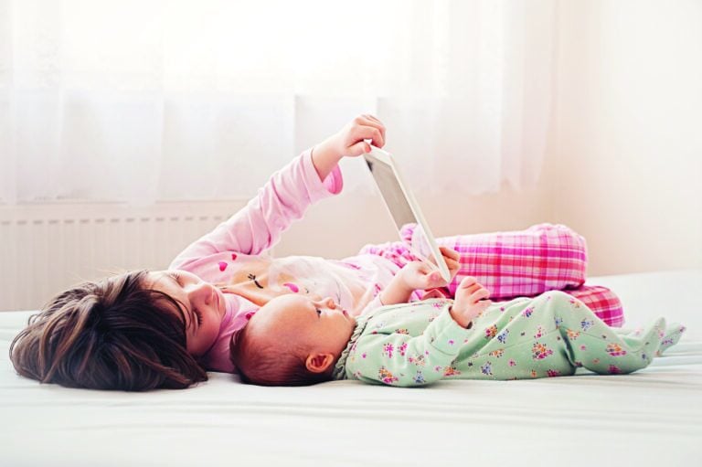 A young girl and her baby sibling are looking at an ipad while resting on a bed